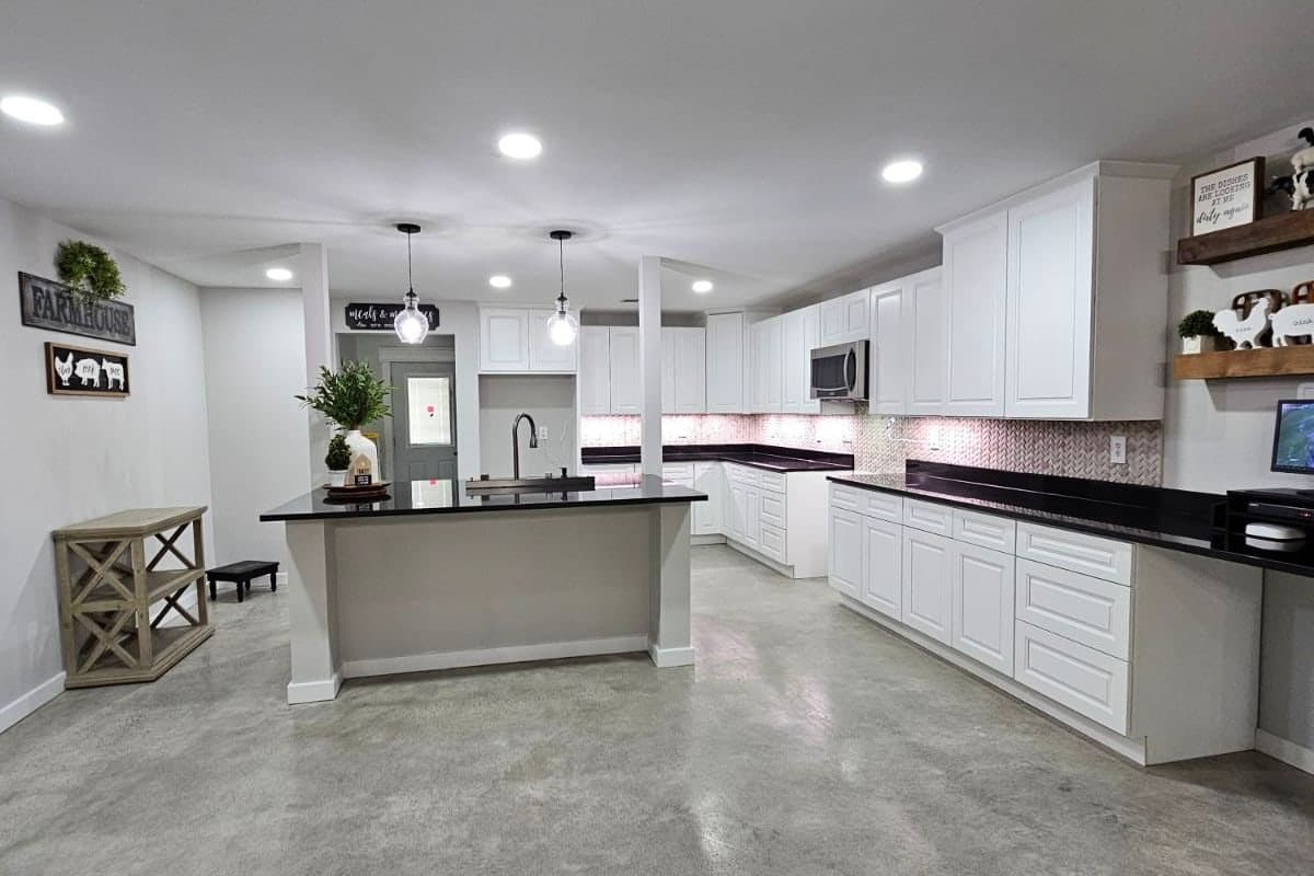 Cocina abierta estilo granja con cubiertas negras sobre gabinetes blancos y una isla central con fregadero, lámparas colgantes y piso de concreto pulido