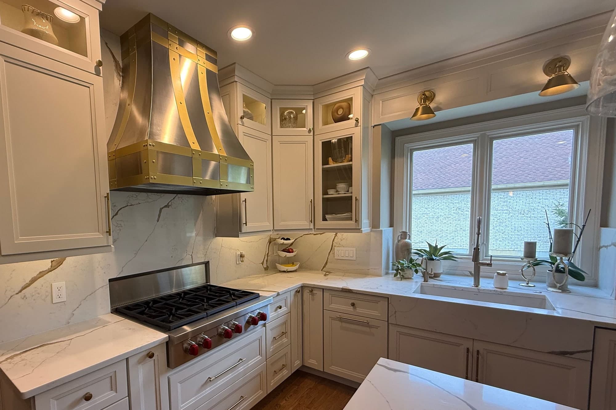 Cocina blanca con cubiertas estilo mármol y salpicadero de la misma piedra hasta el techo, campana de acero y latón, estufa de gas y fregadero bajo cubierta junto a una ventana