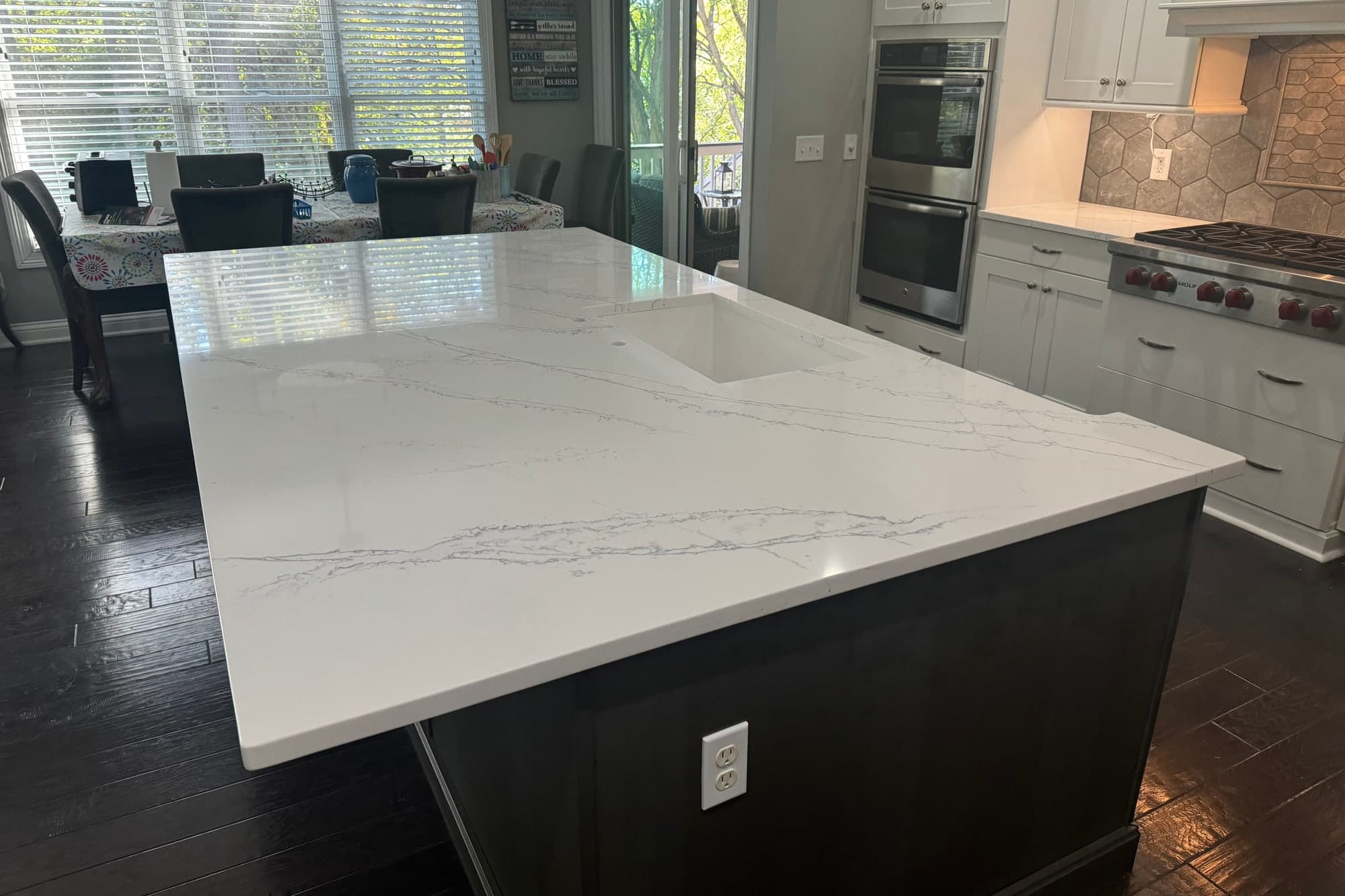 Gran isla de cocina con cubierta de piedra blanca con vetas grises suaves y fregadero bajo cubierta, base oscura y gabinetes blancos con salpicadero de azulejo hexagonal al fondo