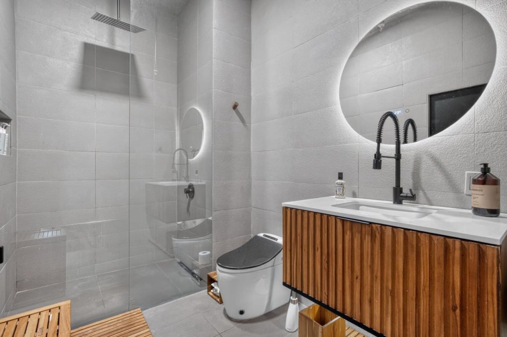Baño a medida con azulejo gris texturizado, mueble flotante de madera acanalada con cubierta blanca, espejo redondo retroiluminado y ducha de vidrio